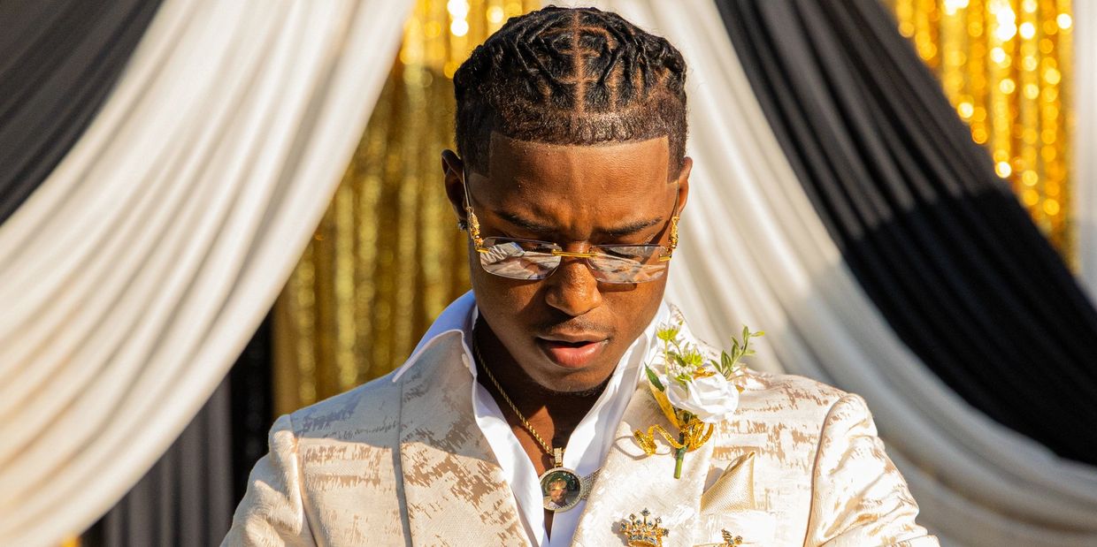 A man in a white suit with gold accents looks down, wearing stylish glasses and a flower boutonniere.