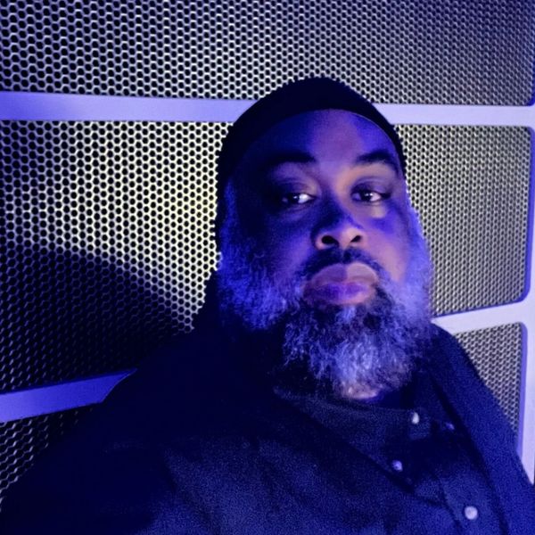 Bearded man illuminated by blue light against a perforated metal background.