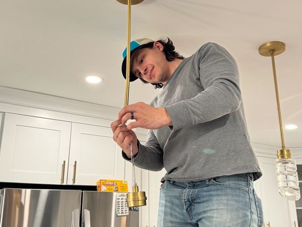Man in a cap installing a pendant light in a modern kitchen.