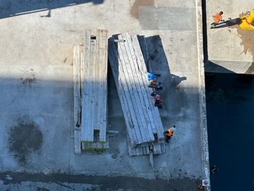 Workers handle long wooden planks on a dock by the water.