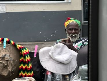Elderly man with colorful headwrap and patterned shirt among hats and baskets.