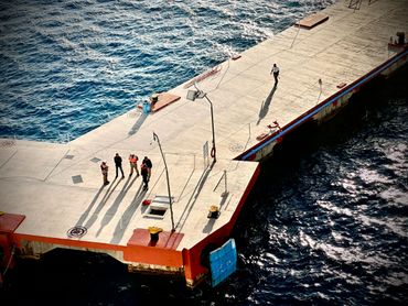 People standing and walking on a sunlit pier by the water.