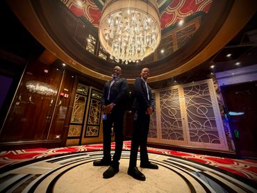 Two men in suits stand back-to-back under a grand chandelier in a luxurious room.