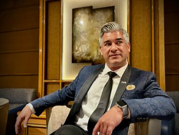 A man in a blue suit sitting confidently on a chair indoors.