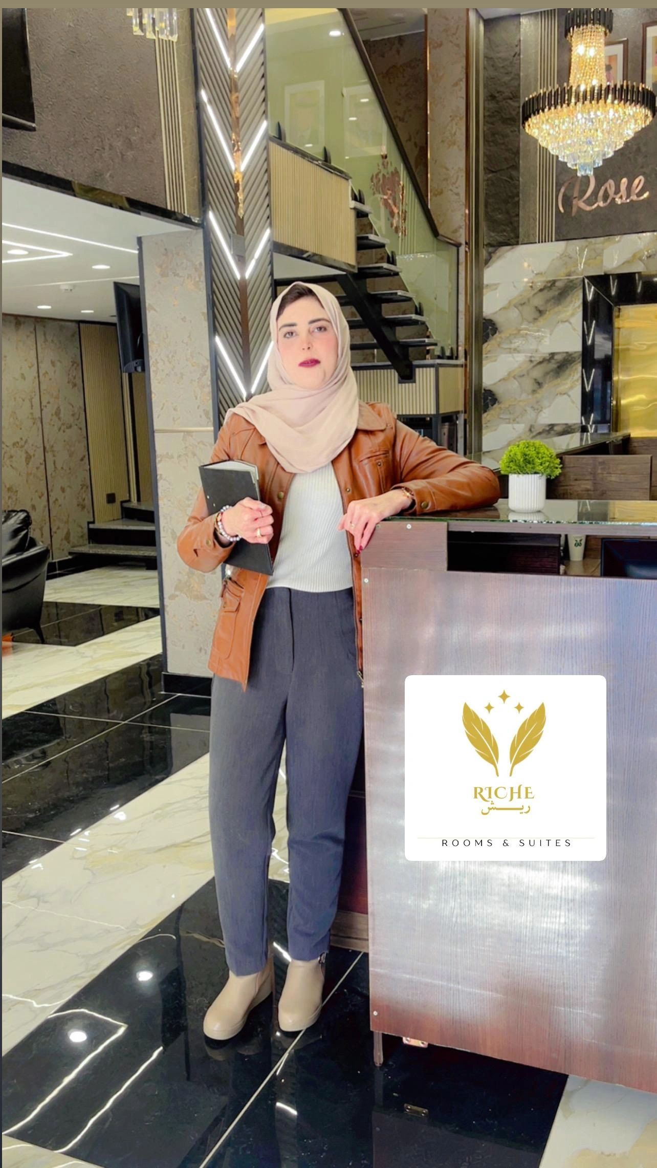 A woman in a hijab and brown jacket stands near a reception desk in a hotel lobby.