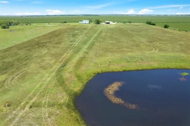 A rural landscape with a pond, grassy fields, and distant buildings under a blue sky.
