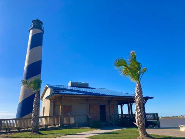 A black and white striped lighthouse beside a small building and palm trees under a clear blue sky.