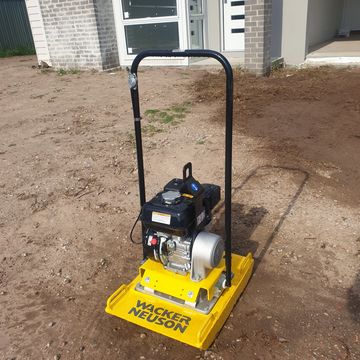Yellow Wacker Neuson plate compactor on dirt near a house under construction.