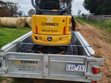 Yellow Corbex Hire excavator loaded on a trailer with rural farm background.