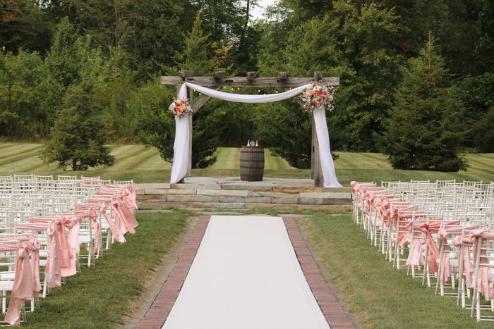 Outdoor wedding ceremony setup with floral arch and pink ribbons on chairs.