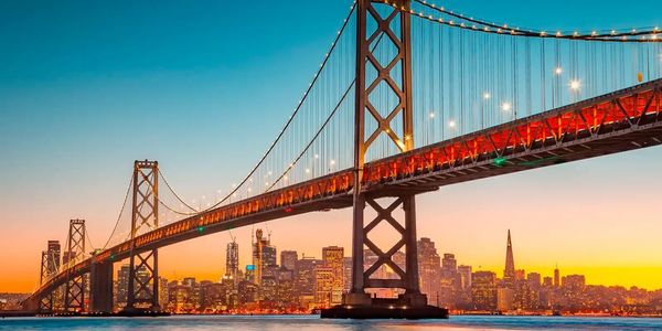 Golden Gate Bridge lit up at sunset with city skyline in background.