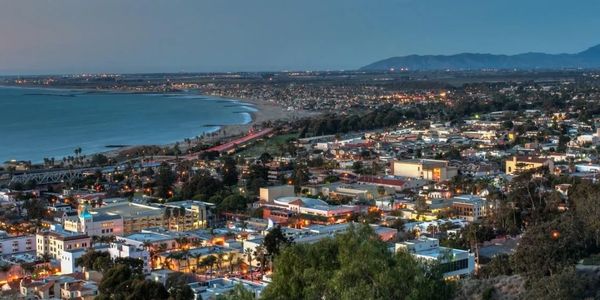 Panoramic evening view of a coastal city with illuminated buildings and distant mountains.