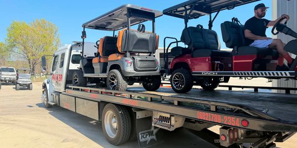 Two golf carts loaded on a flatbed tow truck under clear blue sky.