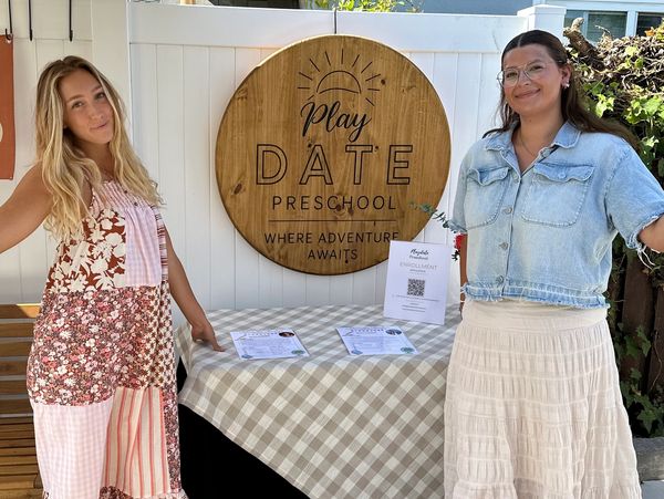 Two women stand by a preschool sign and table.