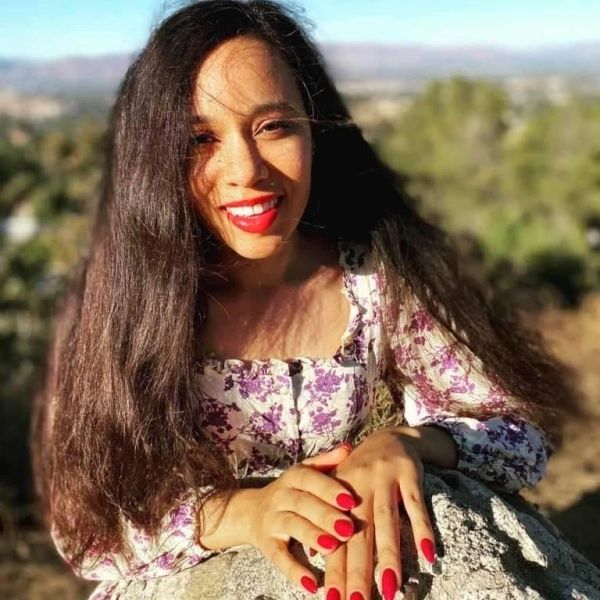 Smiling woman with long hair and red nails in floral top outdoors.