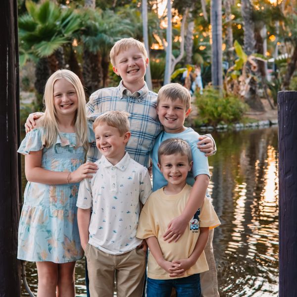 Five smiling children posing by a pond with palm trees.