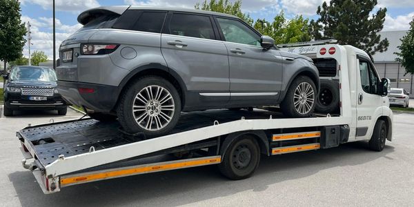 A gray Range Rover being transported on a white flatbed tow truck under a blue sky.