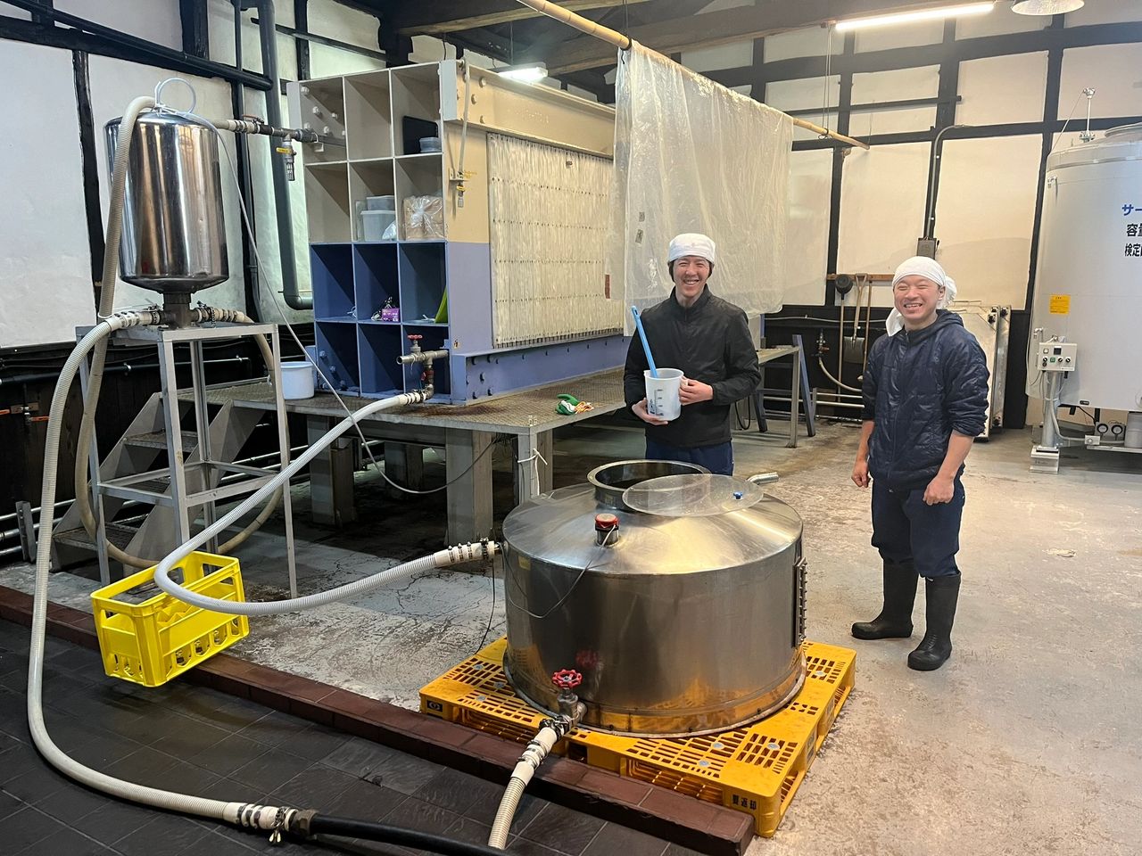 two Japanese kura-bito sake brewer workers sampling Japanese sake after it was pressed by the Yubota pressing machine behind them, holding a container of freshly pressed sake called Shiboritate