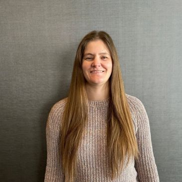 Smiling woman with long hair wearing a knitted sweater against a plain background.