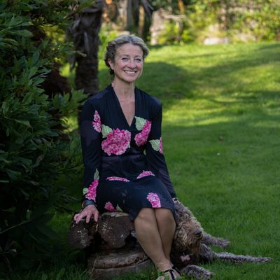 Woman in floral dress sitting on a tree stump with a dog lying beside her in a garden.