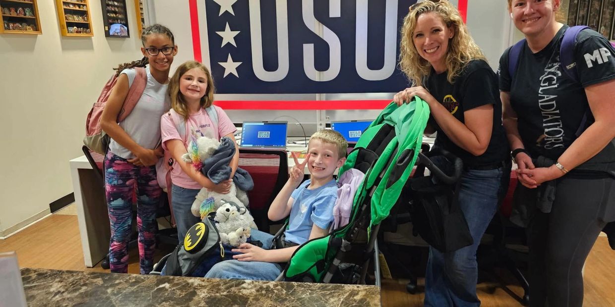 Group of five smiling people posing indoors with USO banner.