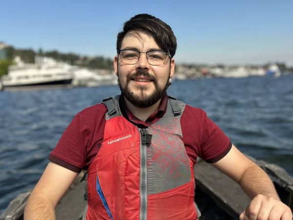 Man in red life jacket rowing a boat on water, smiling at the camera.