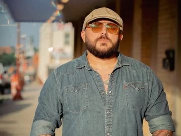 Man in sunglasses and denim shirt standing outdoors in sunlight.
