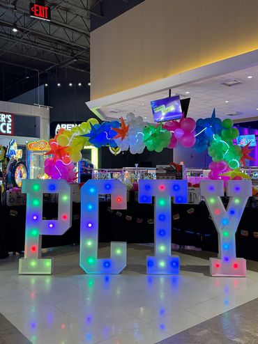 Four illuminated marquee letters PCTY with multicolor lights and neon balloon garland above.