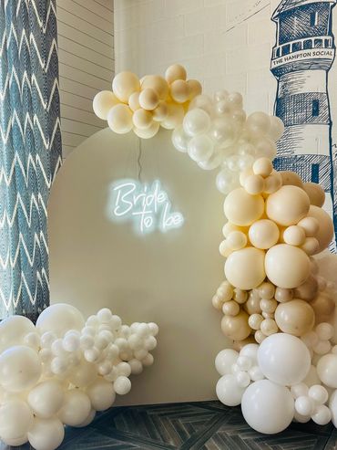 Elegant bridal shower setup with white and beige balloons and a 'Bride to Be' neon sign.