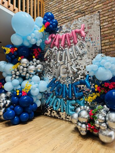 Graduation display with shimmering backdrop, Mediterranean -inspired balloons, and floral accents.