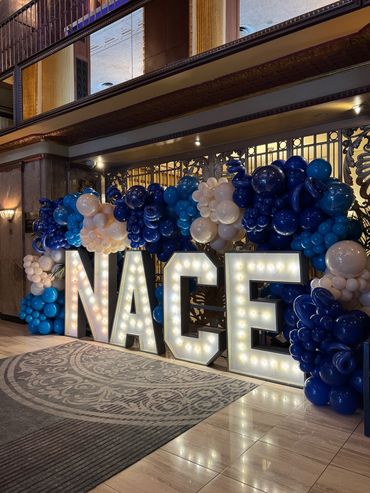 Marquee light up letters spelling "NACE" surrounded by an organic balloon garland. 