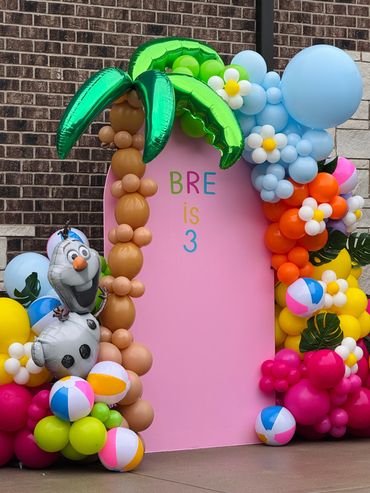 Pink backdrop with balloon palm tree, balloon garland, Olaf, beach balls, and balloon flowers.