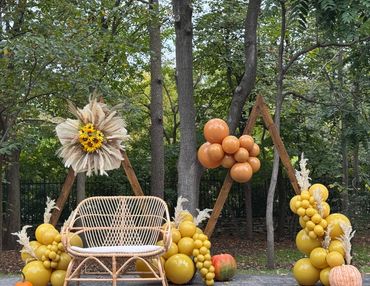 Boho wooden triangle backdrop with balloons, pampas grass, florals, pumpkins, and wicker bench.