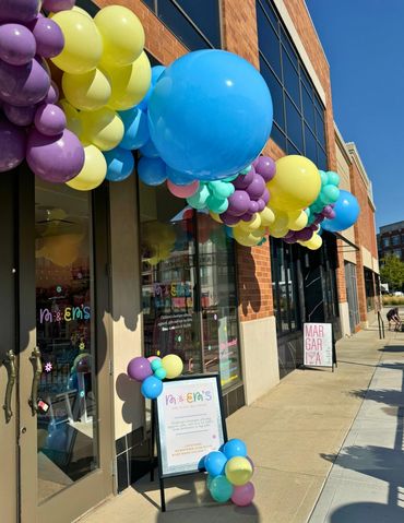 Organic balloon garland in purple, yellow, blue, green, and pink, installed above shop windows.