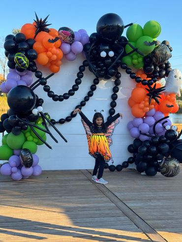 Girl posing in front of a white Halloween backdrop with balloon garland , spiders, and bats.