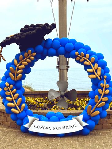 A round graduation balloon hoop in blue, adorned with a graduation hat, gold leaves, and a sign.