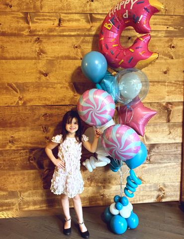 Girl posing with a helium birthday balloon bouquet featuring swirls, candy, stars, and donut balloon