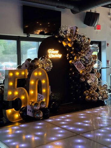 Black and gold birthday backdrop with marquee lights, bubble balloons, casino cards, and neon sign.