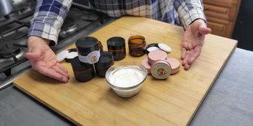 A person presenting various cosmetic jars and a bowl of cream on a wooden board.