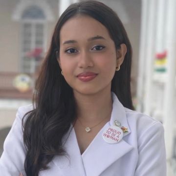 A confident woman wearing a white coat and a pin that says 'Proud to be a woman.'