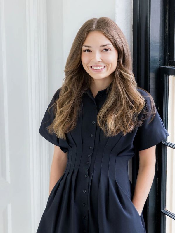 Young woman smiling in black dress with hands in pockets.