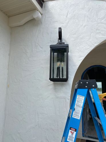 Black outdoor lantern mounted on a white stucco wall with a blue ladder nearby.