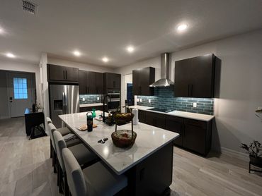 Modern kitchen with dark cabinets, white countertop island, and teal backsplash tiles under warm lighting.