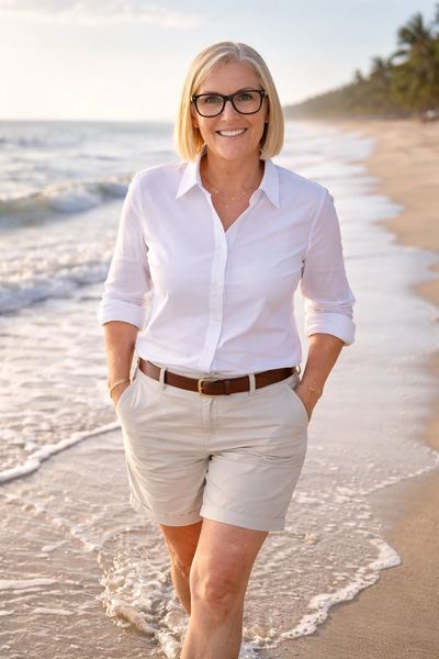 Smiling woman in white shirt and shorts walking on the beach with ocean waves.