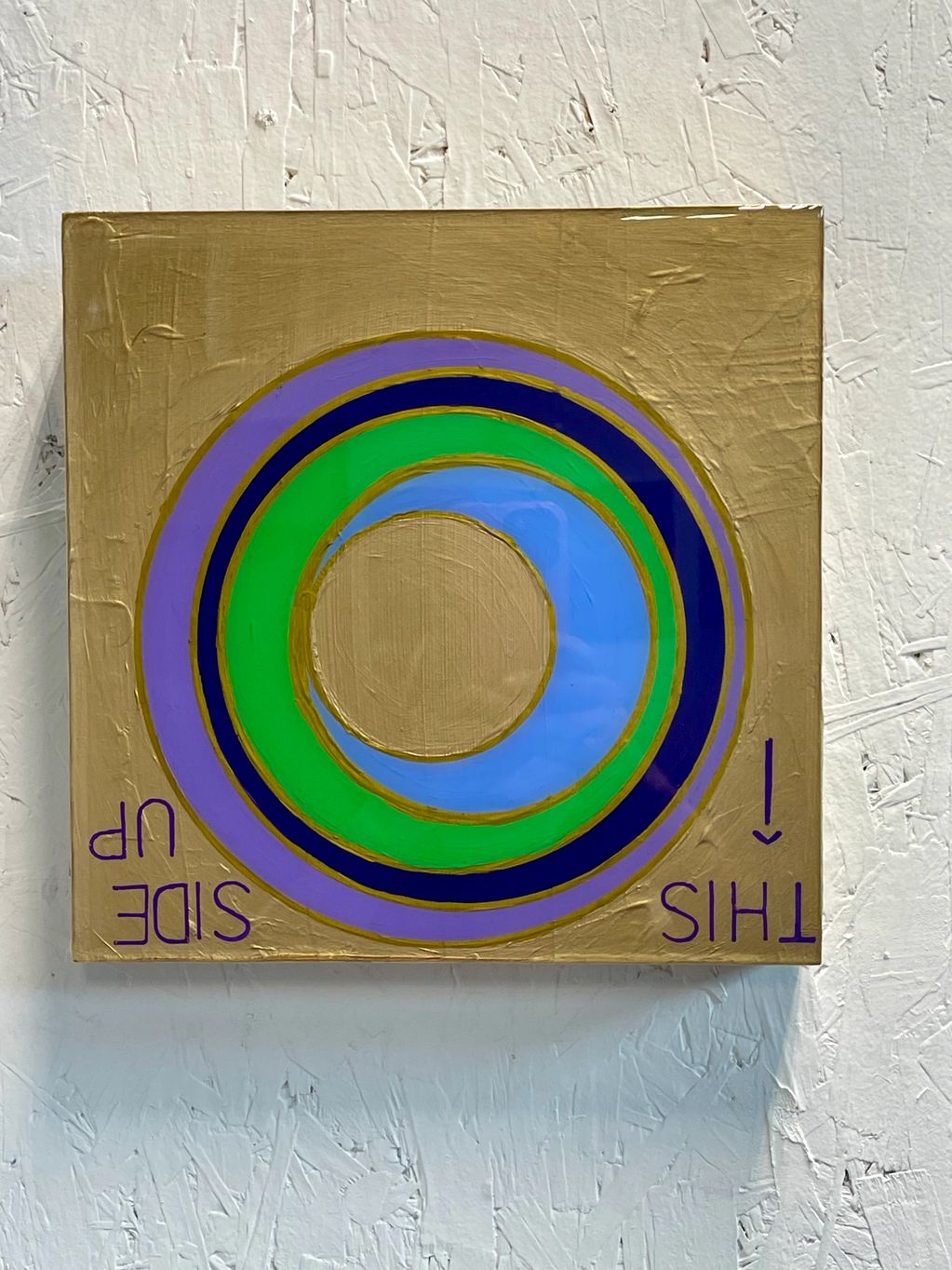 Abstract concentric circles painting with 'THIS SIDE UP' text on gold background.