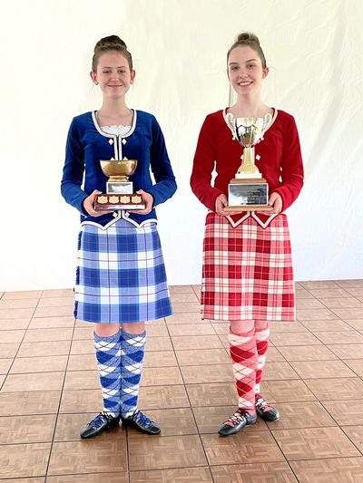 Two girls in Scottish attire hold trophies, one in blue and one in red.