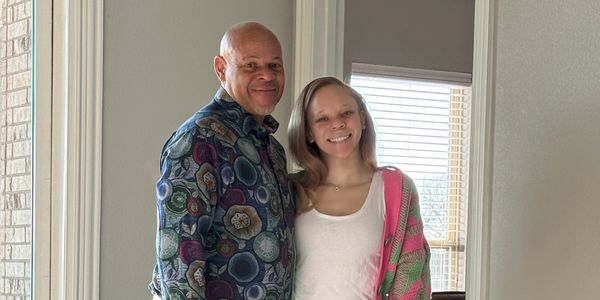 A smiling couple stands together in a well-lit room, dressed casually and colorfully.