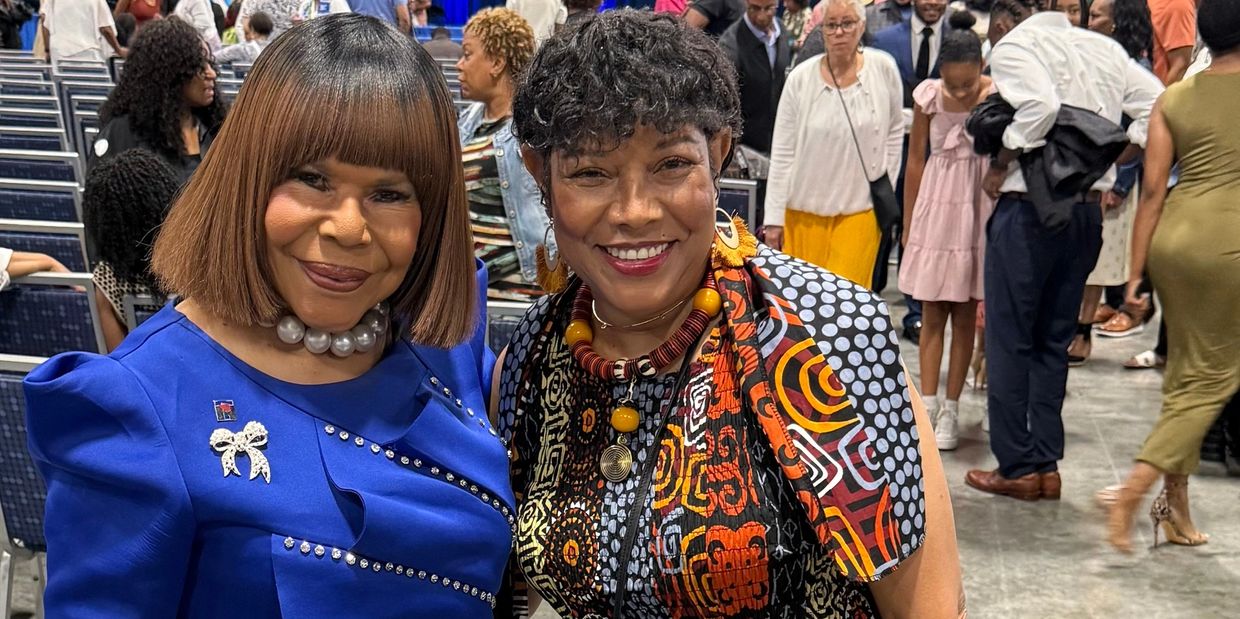 Two stylish women smiling at a social event with a crowd in the background.