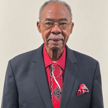 Elderly man in a black suit and red shirt with matching tie and pocket square.