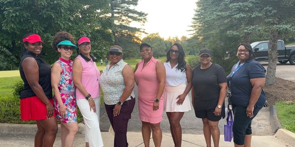 Eight women posing outdoors in golf attire on a sunny day.